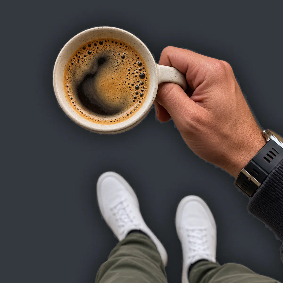 A POV image of a man holding a coffee mug from above, standing in white sneakers and olive trousers.