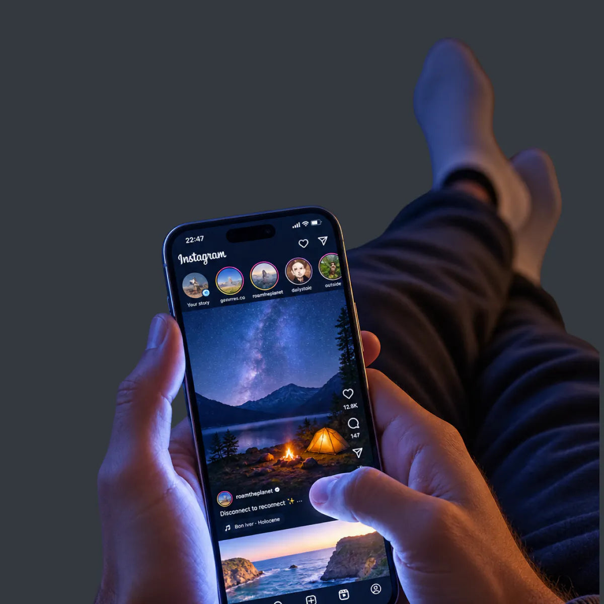 A person holding a smartphone showing the Instagram app with a camping landscape post, with their legs and feet visible in dark socks against a dark background.