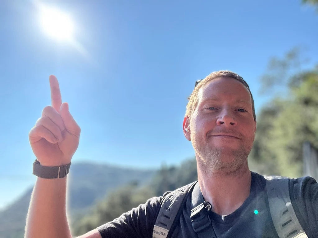 Matt, founder of Gavarres, pointing towards the sun with a clear blue sky and the rolling forested hills of Les Gavarres mountains behind him.